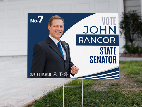  Campaign yard sign on grass in front of a house, featuring a smiling male candidate in a suit with text, along with social media handles and the number 7