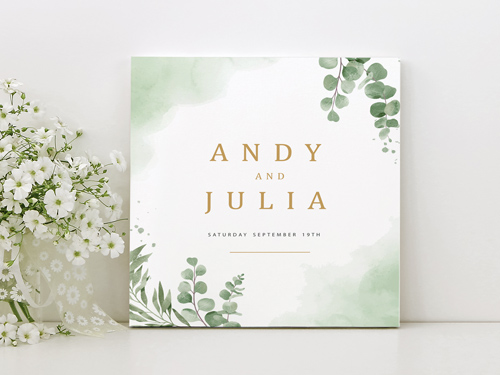 Elegant foam board sign with soft green watercolor foliage and gold typography, announcing the wedding of Andy and Julia, displayed next to delicate white flowers