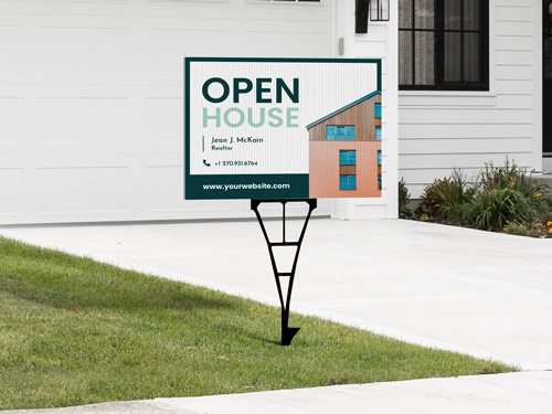 Open house yard sign placed on a green lawn next to a driveway, featuring a modern house graphic, a website URL, supported by a metal stake frame