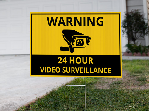 Security yard sign with a yellow and black design placed in mulch near a walkway, featuring a camera icon to indicate that the property is monitored or protected
